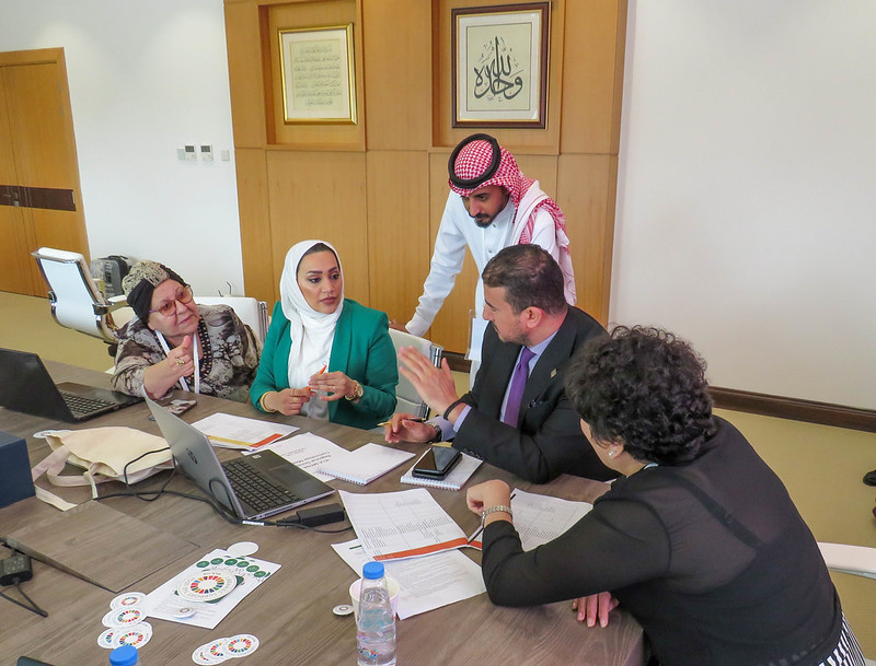 Group of five people discussing at a table, in front of a lap top. From left to right a woman in a black and gold turban-like hat, a woman in a white headscarf and green jacked, a man (standing) in a white tunic and red check headcloth, a man sitting in a dark suit and purple shirt and tie, and a woman, with her back to us, in a black blouse. 