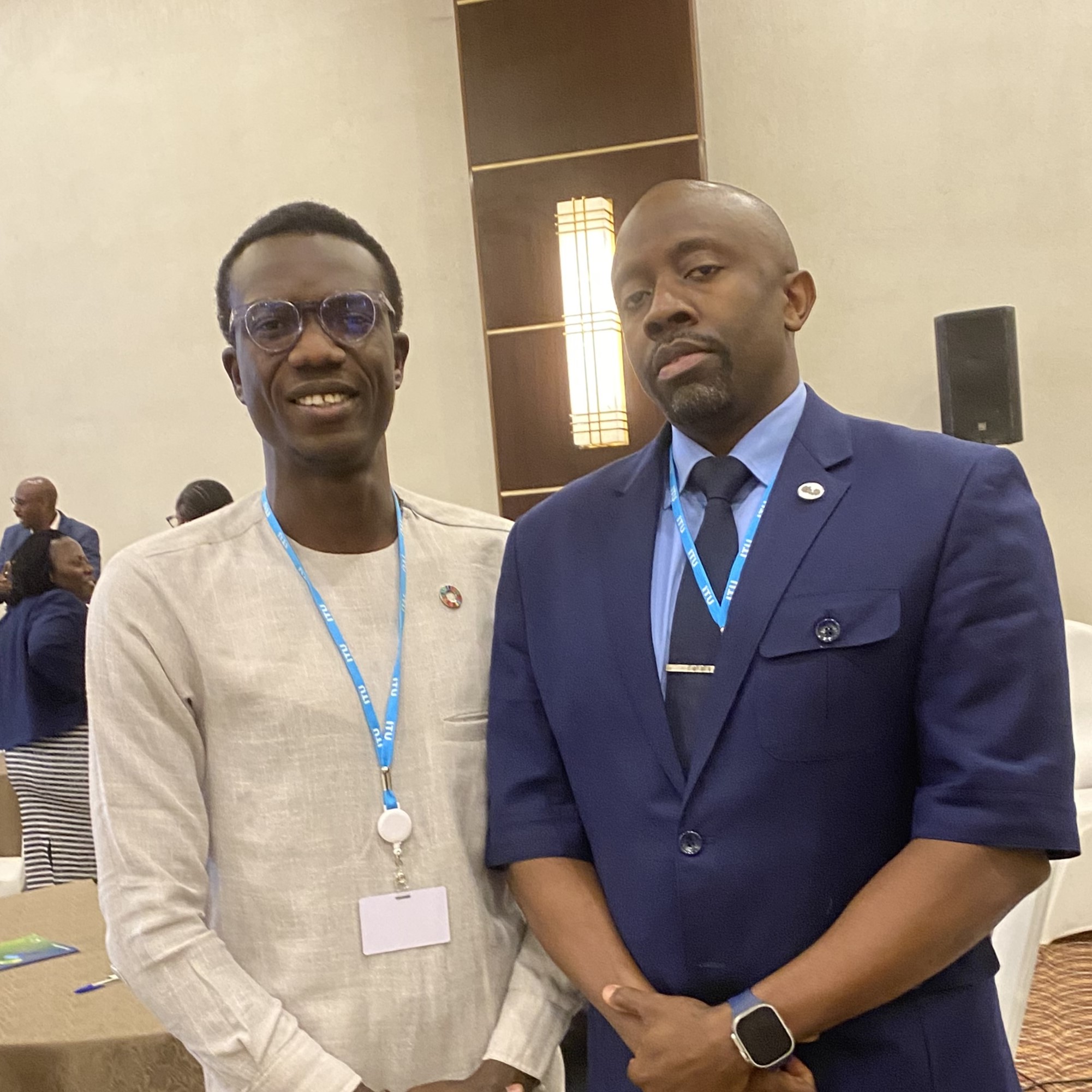 Two African men. On the left a younger one in a beige top with an SDGs badge. On the right, an older one in a blue suit and tie.
