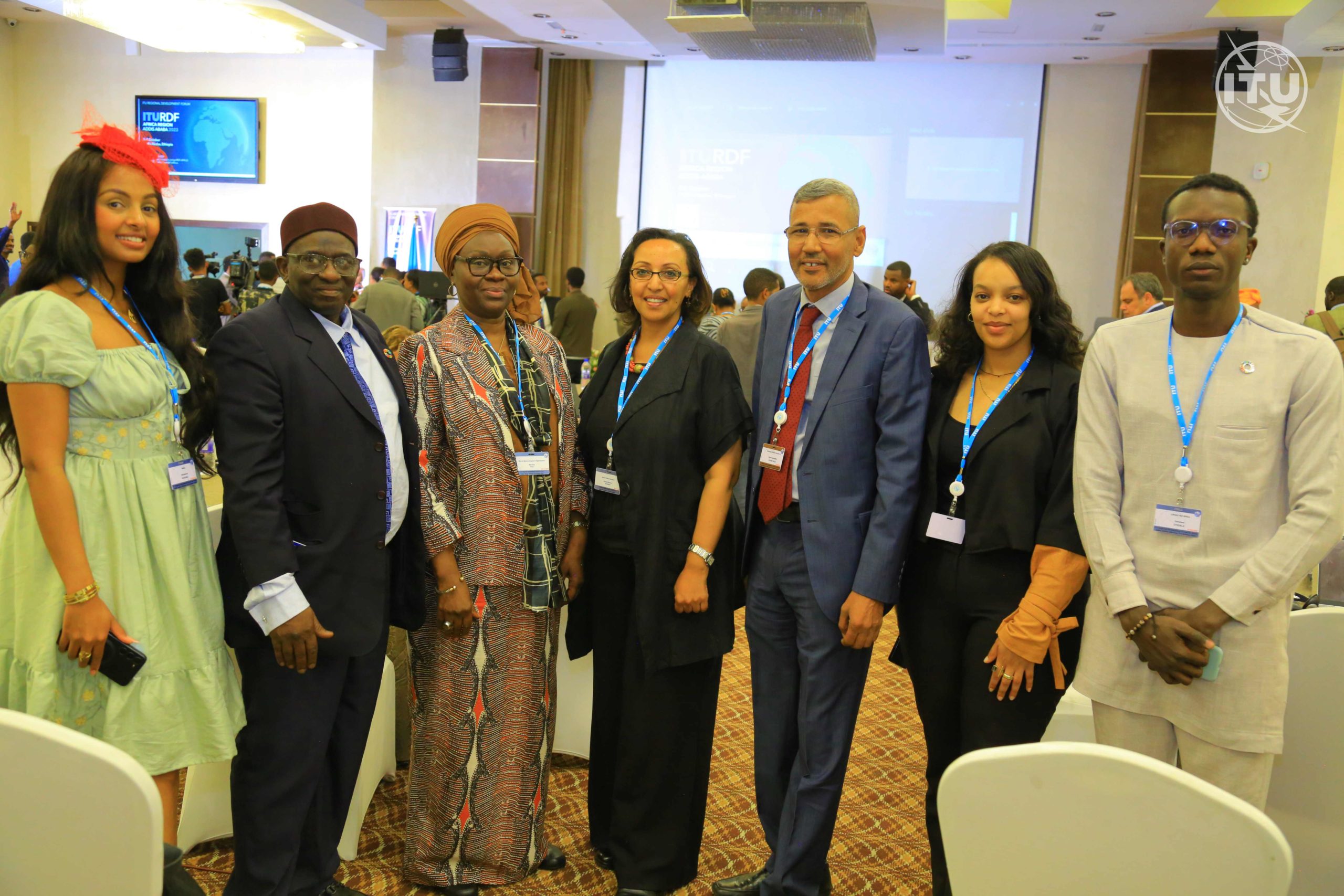 Group of delegates at the International Telecommunications Union Regional Development Forum, Africa. Left to right, a woman in a green dress with a red flower in her hair. A man with a red hat. a woman in a patterned shirt and top, a woman in black, a man in a blue suit with a red tie, a woman in black, and a man in a beige top and trousers. 