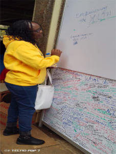 African woman with a yellow hoodie, blue jeans, mid-length black hair and glasses, writing 'Libraries Change Lives' on a whiteboard