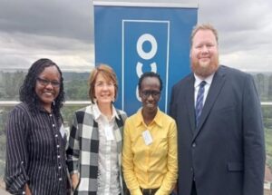 Four people (left to right) an African woman with a black striped shirt, mid-length hair and glasses. A white woman with shortish hair, and a big check jacket over a white shirt. An African woman with a yellow shirt, short hair and glasses, and a man with a beard, suit and tie